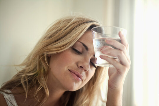 Young woman holding glass of water against her forehead at home