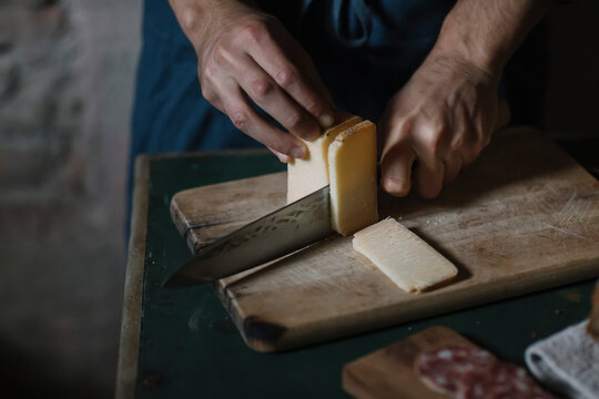 Hands Of Man Cutting Artisanal Cheese Slices On Board At Table