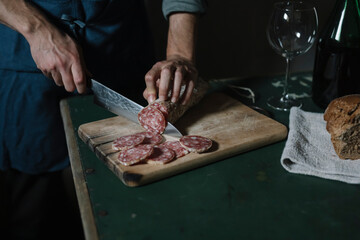 Hands of man cutting salami slices on board at table