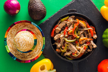Frying pan with tasty beef Fajita, vegetables and sombrero hat on color background
