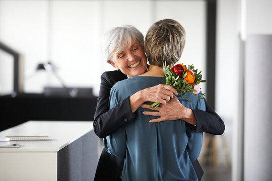 Happy senior businesswoman with bunch of flowers hugging colleague in office