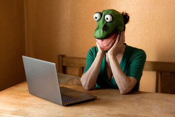Playful mature woman wearing frog mask with laptop on table sitting at home during curfew