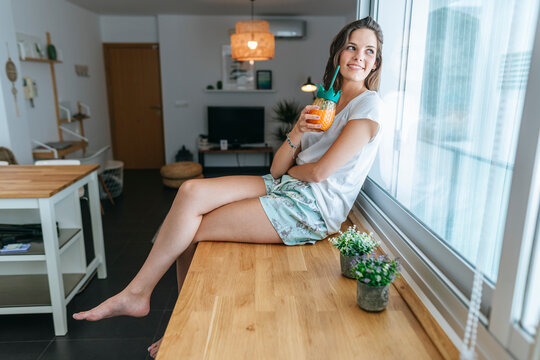 Smiling Young Woman Sitting On Kitchen Counter With A Drink Looking Out Of Window