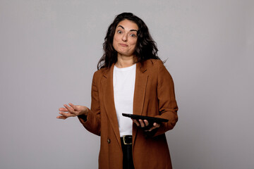 Confused woman with digital tablet gesturing against gray background