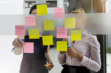 Female colleagues discussing over adhesive notes stuck on glass wall in office
