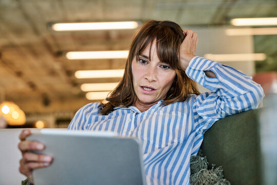 Woman with shocked expression looking at digital tablet while sitting on sofa at home