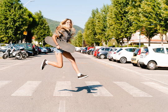 Cheerful young woman holding skateboard while jumping on zebra crossing in city