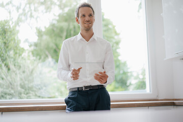 Businessman standing at the kitchen window, using glass touch screen, smiling