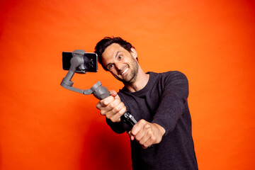 Cheerful man taking selfie while holding gimbal with smart phone against orange background