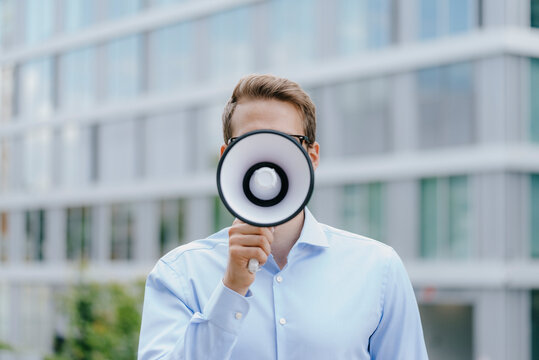 Young businessman standing in front of modern office building, using megaphone