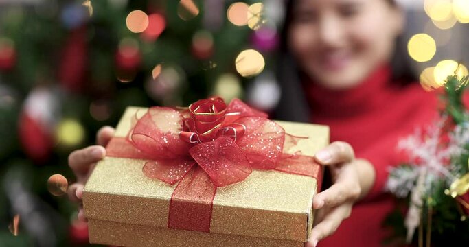 Young teen cute Chinese girl holding present box offer it gives to camera in room with Christmas tree and ornaments, boke lights in background. Idea of Xmas blessing new normal life.