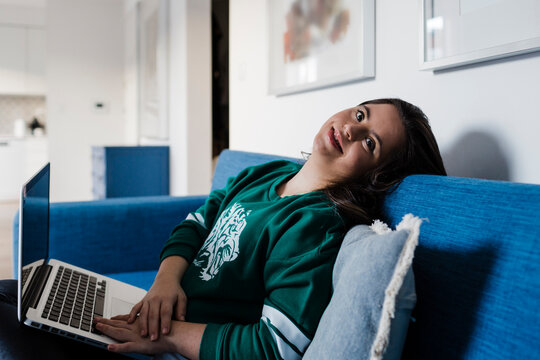Happy young woman with down syndrome using laptop while relaxing on sofa at home