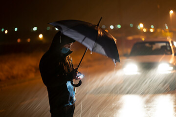 Young man with umbrella and face mask using mobile phone while standing against breakdown car during rainfall