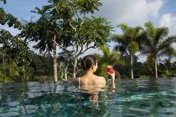 Woman drinking cocktail on infinity pool with stunning tropical view. Back view