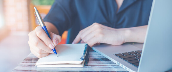 Business woman hand writing on a notepad with a pen and use a laptop computer in the office.