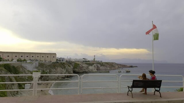 Mother and child sit on bench under Italian waving flag overlooking Terrasini coastline and cove, Sicily. Italy