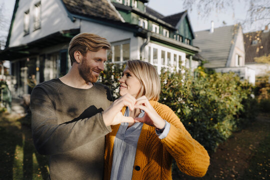 Smiling couple standing in front of their home shaping a heart with their hands