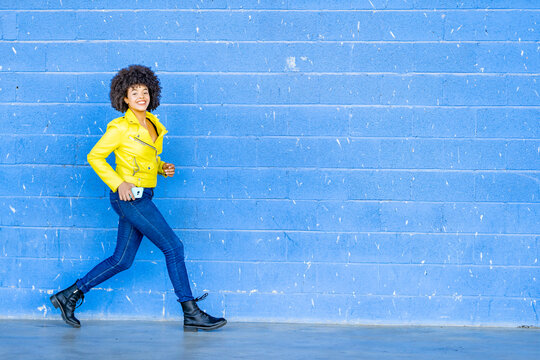 Happy woman in yellow leather jacket holding mobile phone while walking against blue wall