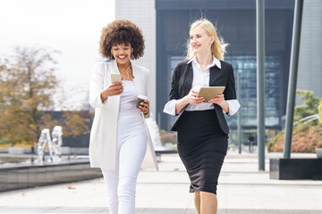 Smiling businesswomen walking with smart phone and digital tablet on footpath