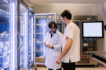 Male pharmacists taking inventory while standing by illuminated refrigerator in hospital