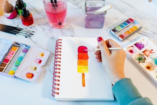 Woman painting popsickle ice cream with watercolor in book on table