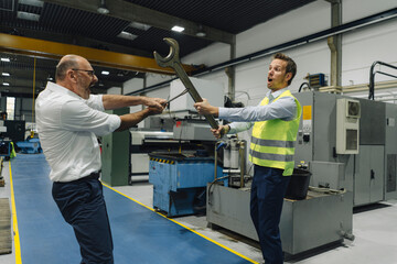 Businessman and man in reflective vest playfighting with large wrenches in a factory