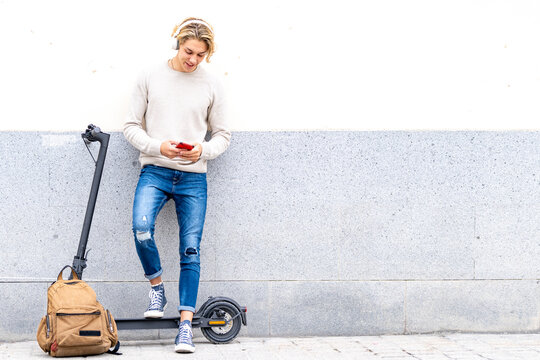 Man with backpack and electric push scooter using mobile phone while standing against wall