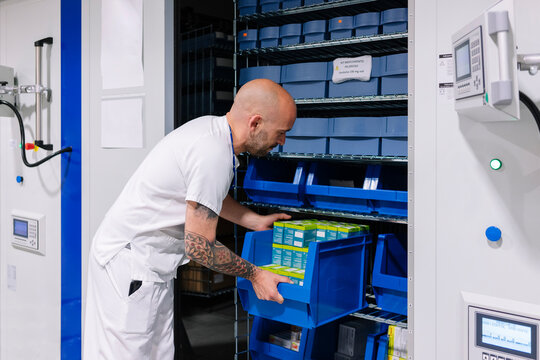 Bald male pharmacist placing trays of medicines in shelf at hospital