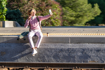 Smiling man gesturing peace sign while taking selfie sitting with longboard on station platform