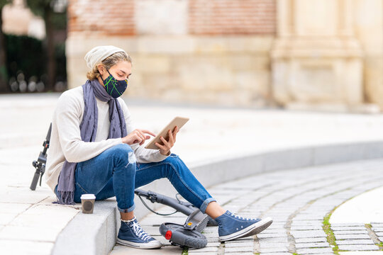 Young man with electric push scooter and face mask using digital tablet while sitting on footpath in city