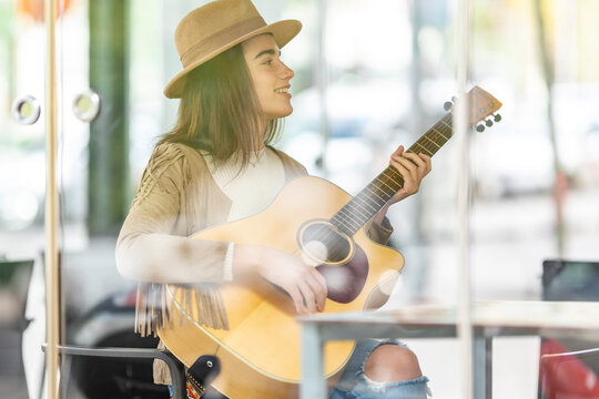 Smiling young female musician playing guitar seen through glass at cafe