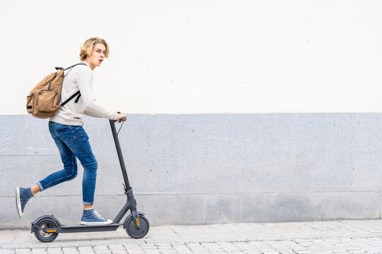 Man with backpack riding electric push scooter on footpath against wall
