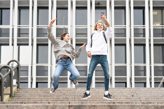 Carefree friends with taking selfie through smart phone while jumping over steps against building