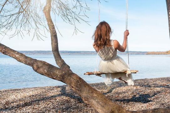 Rear view of woman on swing by tree while looking at view from lakeshore