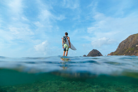 Surfer holding his surf board, standing on stone, Sumbawa island, Indonesia, over-under image