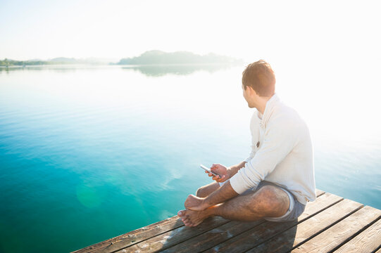 Young man with smartphone sitting on jetty at lake by sunrise looking at distance