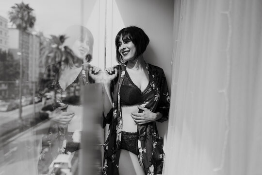 Smiling woman in lingerie looking through window while standing at balcony