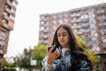 Low angle view of teenage girl using smart phone while standing against chainlink fence