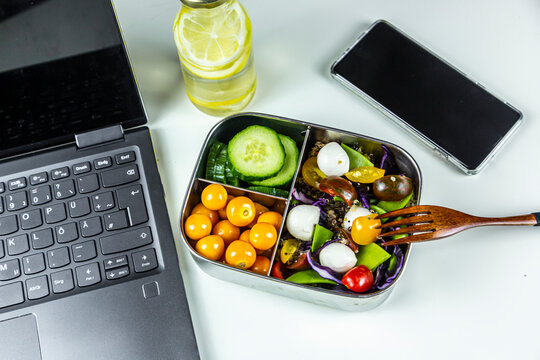 Laptop, smart phone, bottle of lemonade and lunchbox with cucumber slices, winter cherries and quinoa salad (quinoa, cherry tomato, red cabbage, sugar snap peas and mozzarella balls)