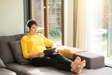 Portrait of woman with digital tablet sitting on the couch at home listening music with headphones