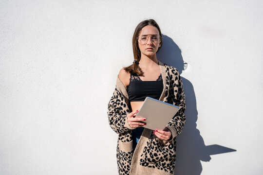 Sunlight falling on stylish teenage girl holding digital tablet while standing against white wall at home