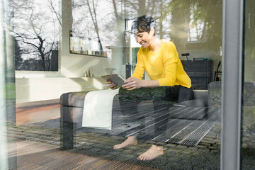 Laughing woman sitting behind windowpane on couch in the living room looking at digital tablet