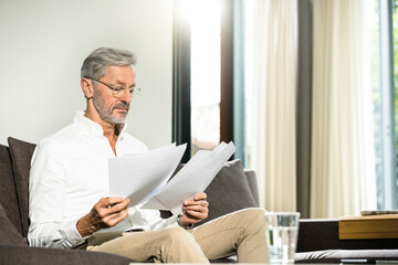Senior man with grey hair in modern design living room sitting on couch working on papers in home office