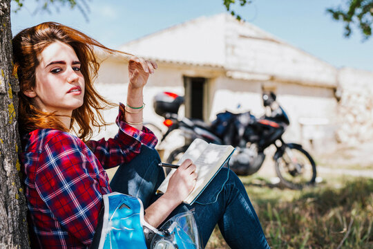 Portrait of redheaded motorcyclist leaning against tree trunk taking notes, Andalusia, Spain