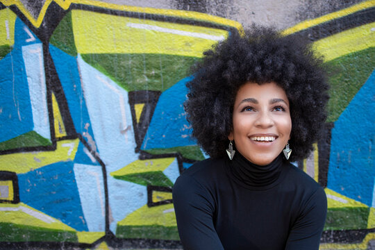 Happy afro young woman looking up while sitting against graffiti wall