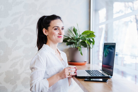 Female Entrepreneur holding cup of coffee while looking through window in coffe shop