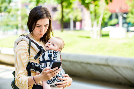 Mother with her baby boy in baby carrier using smartphone in city