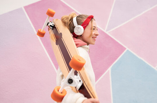 Smiling woman with headphones carrying skateboard on shoulders while standing against colorful wall