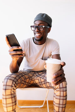Smiling young man using mobile phone while sitting with coffee cup against white wall at home