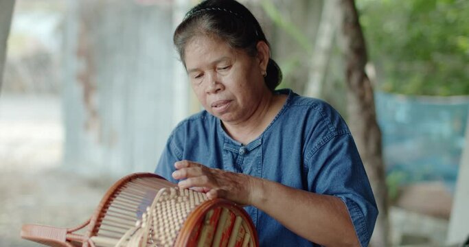A smiling Thai female farmer weave baskets in traditional Thai art patterns to sell as extra income in countryside of Thailand.